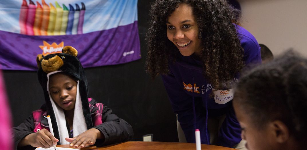 Two kids sit at a table to color while an adult looks on