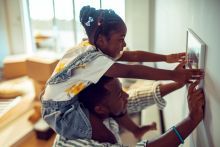 A girl sits atop a man's shoulders as they hang a photo on the wall