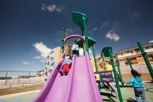 Child stands at the top of the slides and watches another child slide down. A third child runs by the slides.