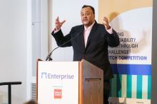 Man with short dark hair wearing a white collar dress shirt and black suit jacket stands behind a podium, with his hands wide open, and a colorful banner behind him.