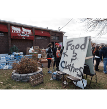 A group of people stand by boxes in front of a building with a red awning that reads Catering Hall and a sign that reads Food and Clothes 