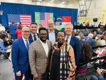 Group of six people pose for photo with flags and signage in the background