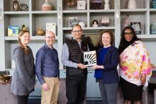 Group of five people stand in front of shelving with two center people holding a plaque.