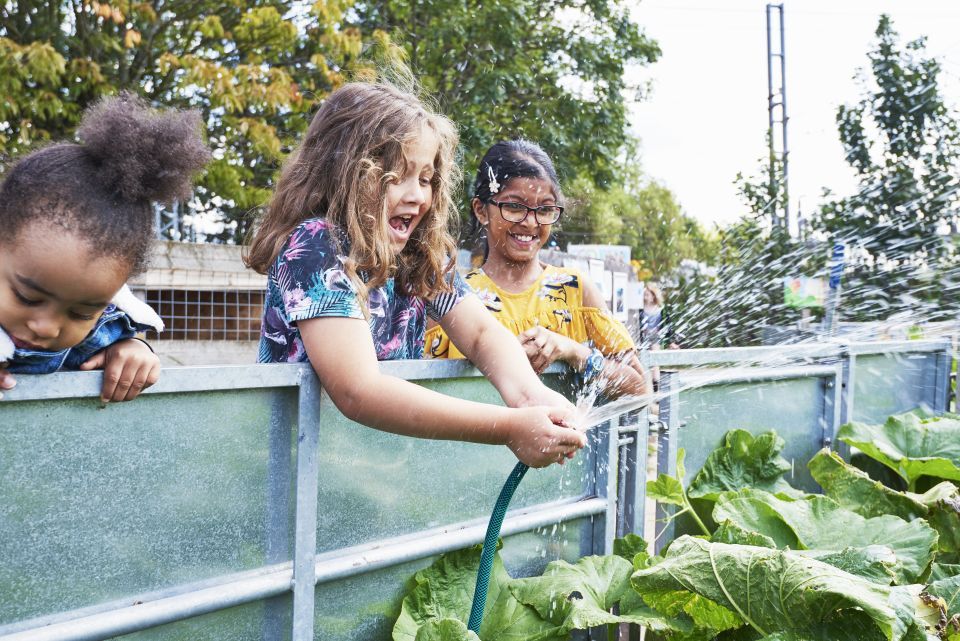 A young girl in a blue top delights in watering a garden with a hose. Two more young girls stand to her right and left watching.