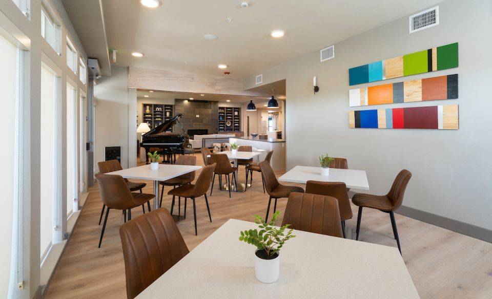 A common area with white cafe tables and brown chairs and brightly colored abstract art on the wall, with a sitting area featuring a baby grand piano in the background.