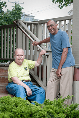 An older adult stands at the bottom of the staircase to talk with an older adult sitting on the stairs