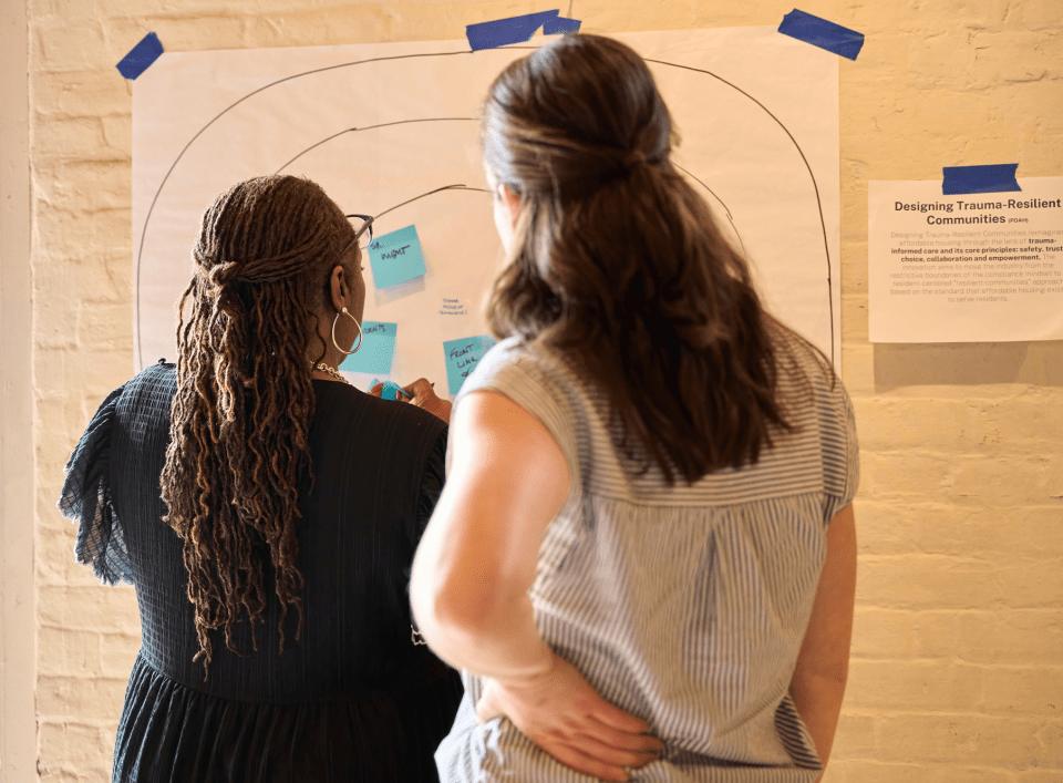 Two people at a poster taped on a wall with a description beside it that reads "Designing Trauma-Resilient Communities"