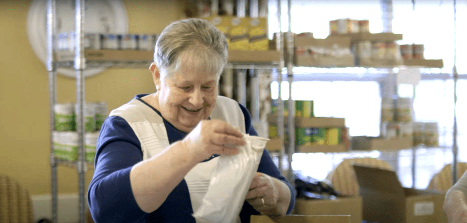 A woman looks at a small package with shelves of food behind her