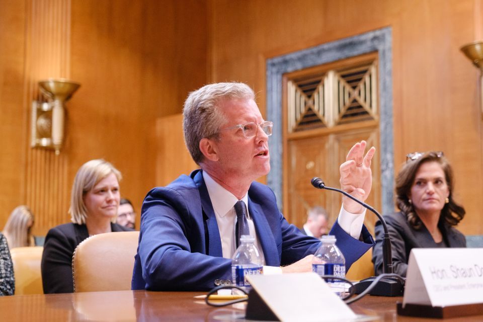 Shaun Donovan, CEO and president at Enterprise, sits in a blue suit at a table and speaks into a microphone with other people in the room