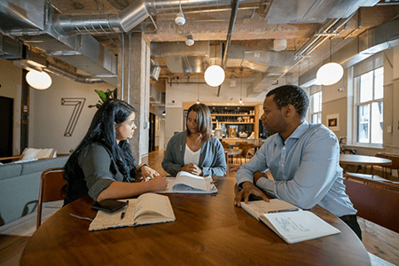 Three people sit and talk at a table with notebooks and pens in a large workspace