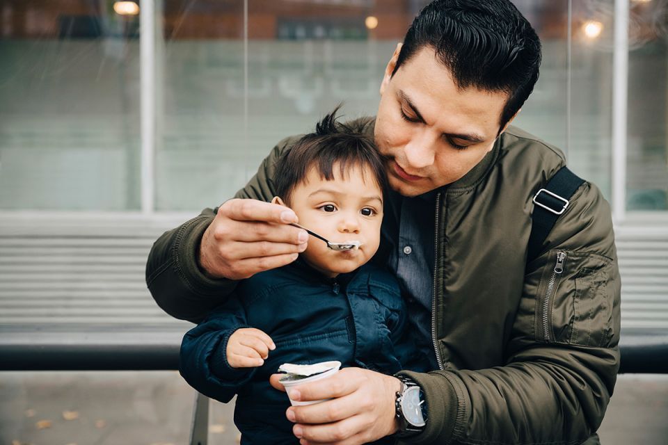 Father sits and feeds his young child who is sitting on his lap 