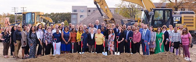 A large group of people standing at a construction site with construction equipment. The first row of people is holding shovels.