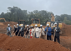 A group of people standing in a row holding shovels in the dirt with three construction vehicles behind them