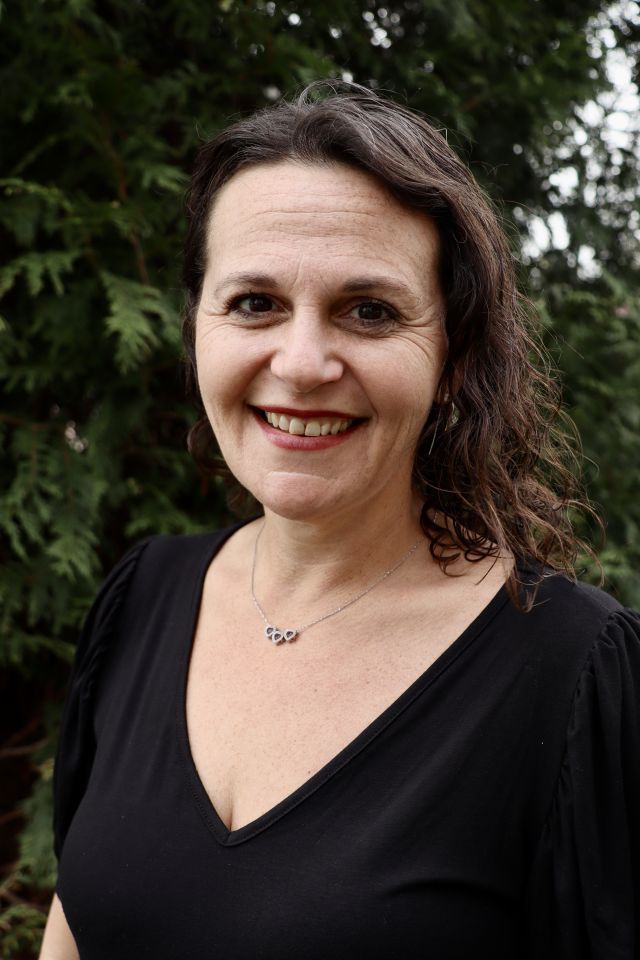 Portrait photo of a woman smiling with brown shoulder-length hair, wearing a black blouse. There is a green foliage in the background.