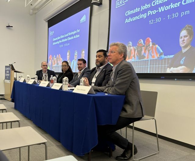 Row of five people speak to an audience from a long table with a blue tablecloth and name tags and two large screens in the background.