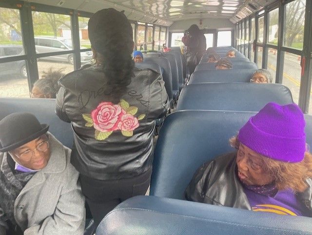 People seated on a bus with blue seats; one person stands in the aisle with their back to the bus.