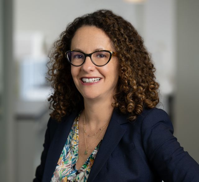 Person with should-length dark curly hair and glasses wearing a dark blazer and multicolored blouse