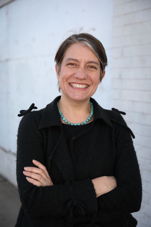 Person with short dark hair in a black shirt stands smiling with her arms crossed