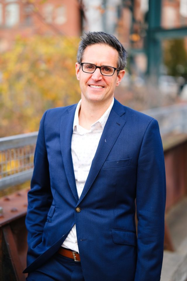 Person with glasses and short black hair stands outside with fall foliage in the background