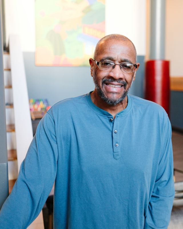 Person with glasses wearing blue Henley shirt stands with artwork in the background