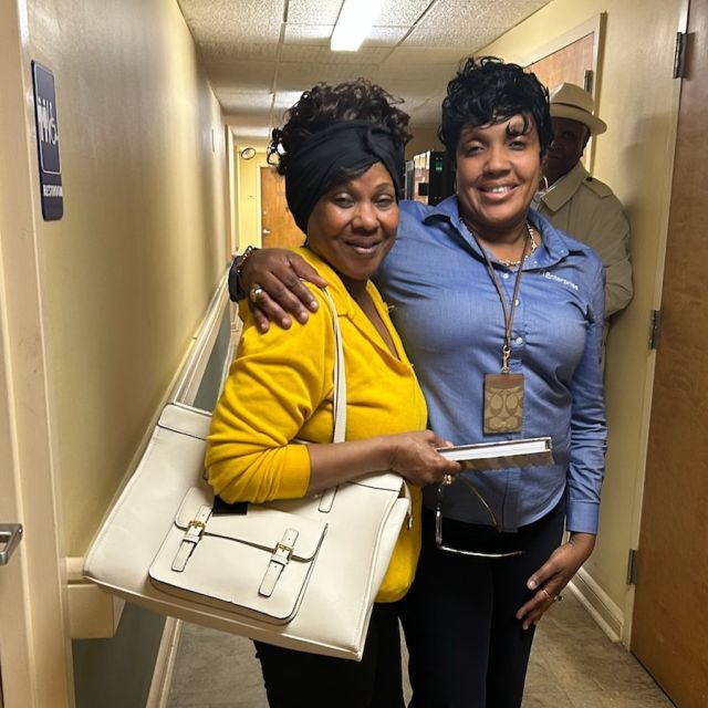 Two people stand next to each in a hallway, with the person on the right wearing a blue shirt and wrapping her arm around the shoulder of the person in a yellow shirt who carries a large white tote purse.