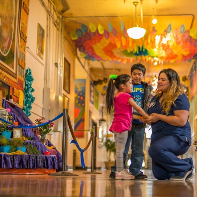 A woman kneels while holding the hands of a young girl as a boy looks on in a decorated hallway