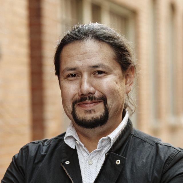 Person with dark hair pulled back and a beard and moustache stands in front of a brick building