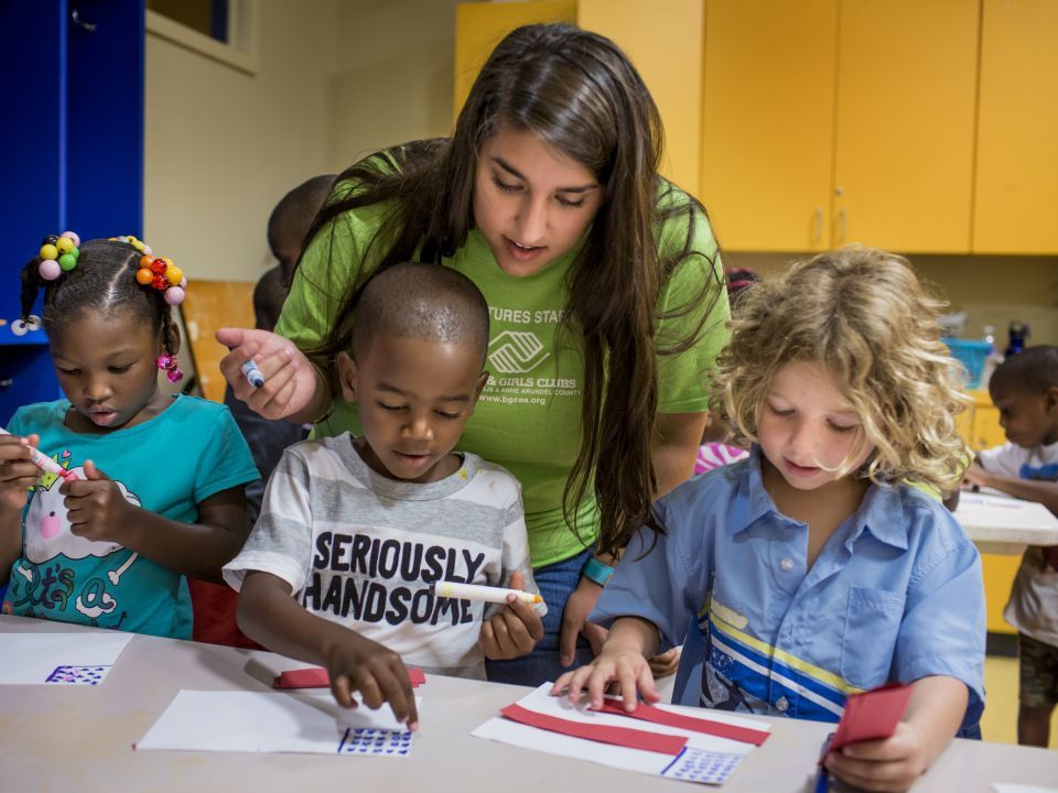 A teacher helps three children sitting at a table with their schoolwork